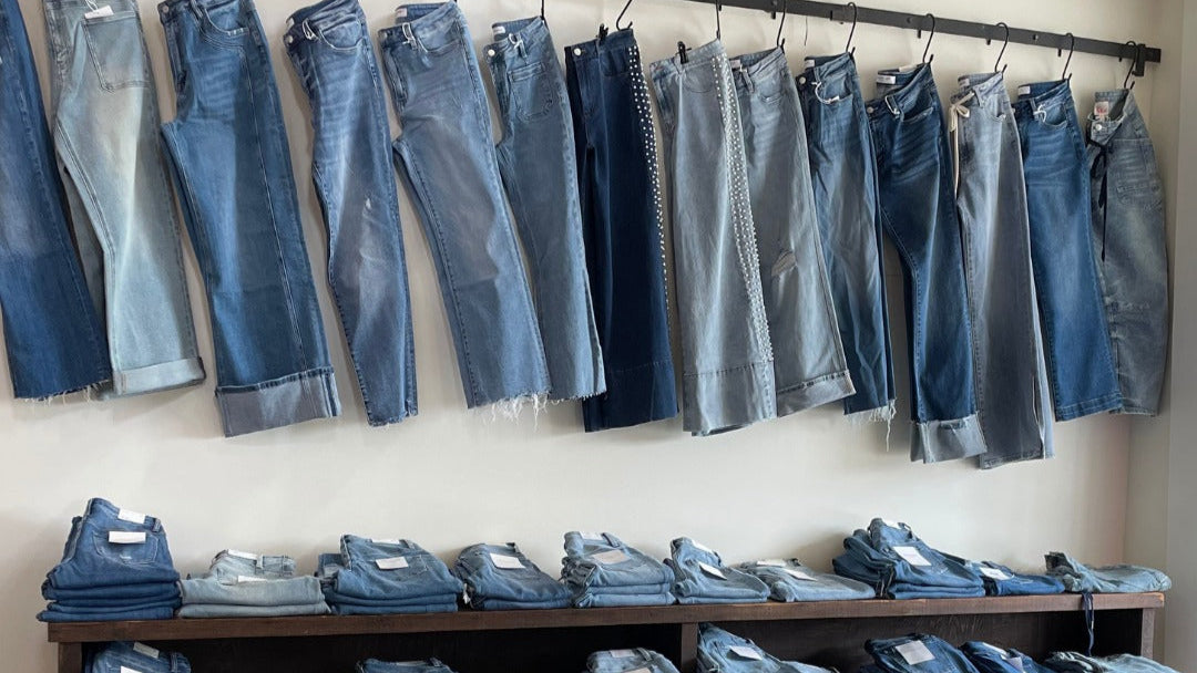 Row of blue jeans hanging on a rack with folded jeans displayed below. - Vervet Jeans - Stretch - Curvy 